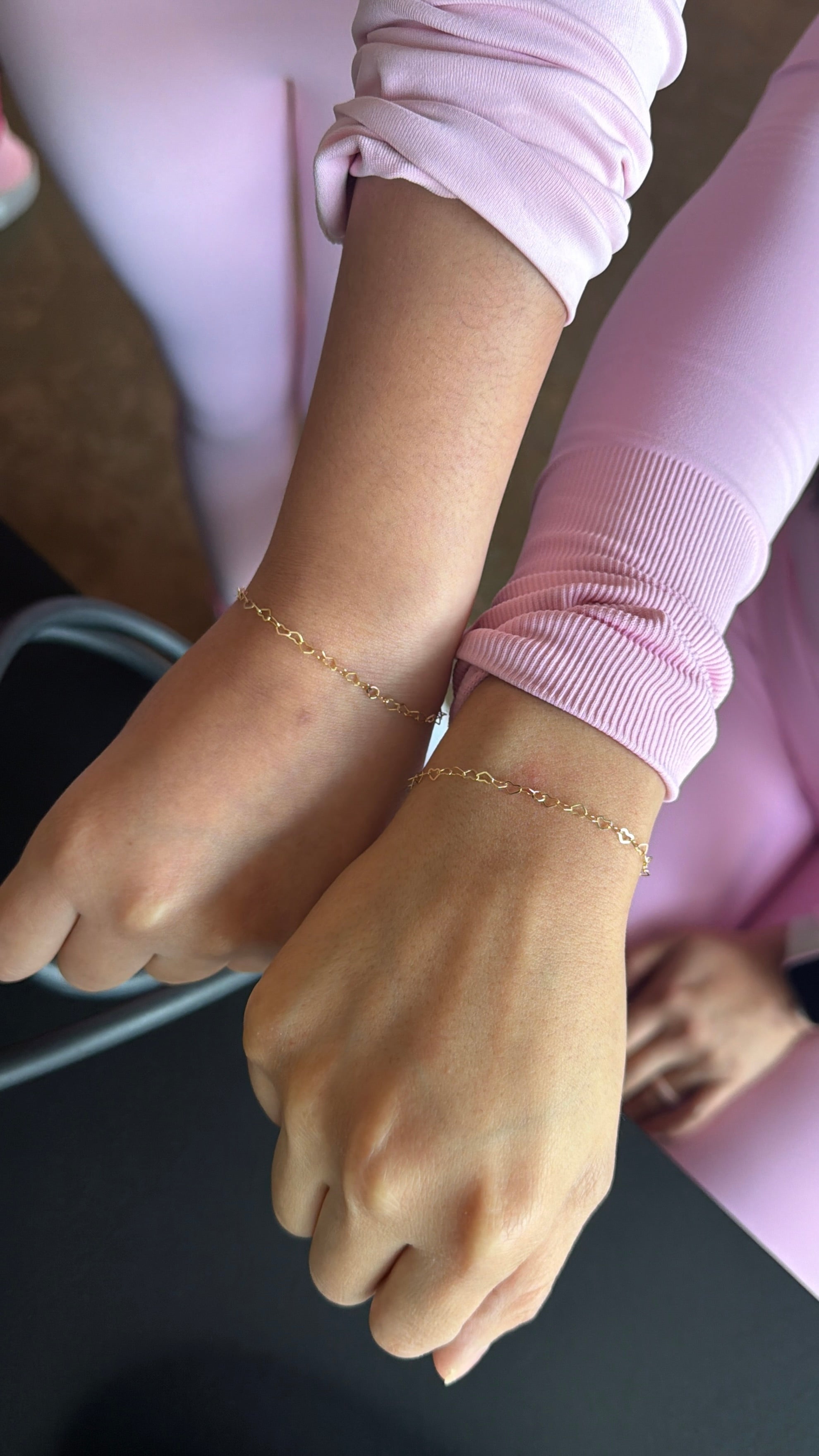 Close-up of two hands wearing gold heart bracelets on a neutral background
