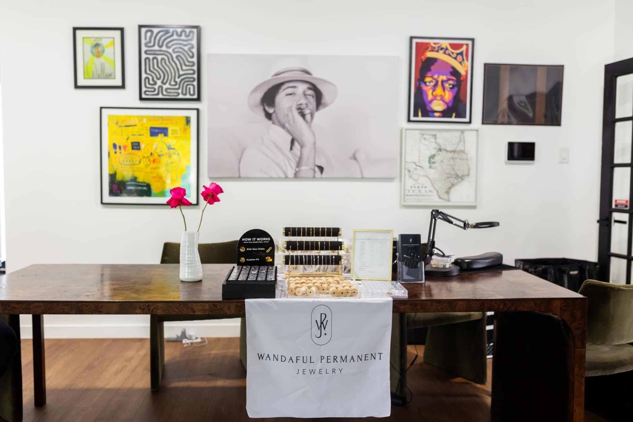 Office space with a desk featuring 'Wandful Permanent Jewelry' and various framed artworks on the wall.